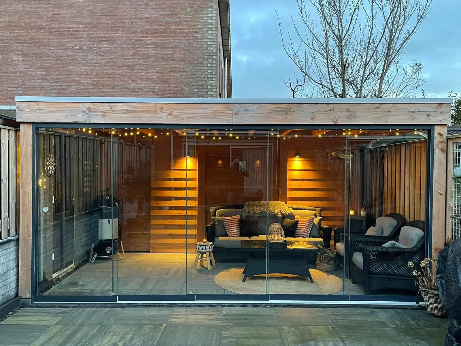 Verlichte glazen veranda met houten achterwand en een loungehoek