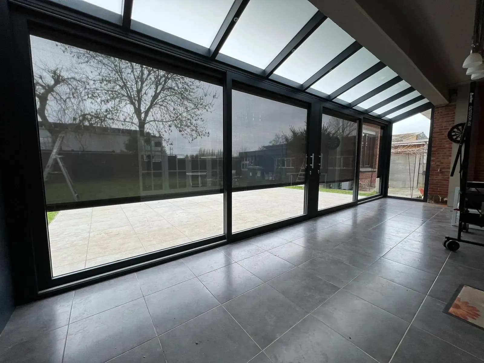 Zicht vanuit een veranda door grote glazen schuifwanden op de tuin, met een lichtdoorlatend dak erboven en een tegelvloer