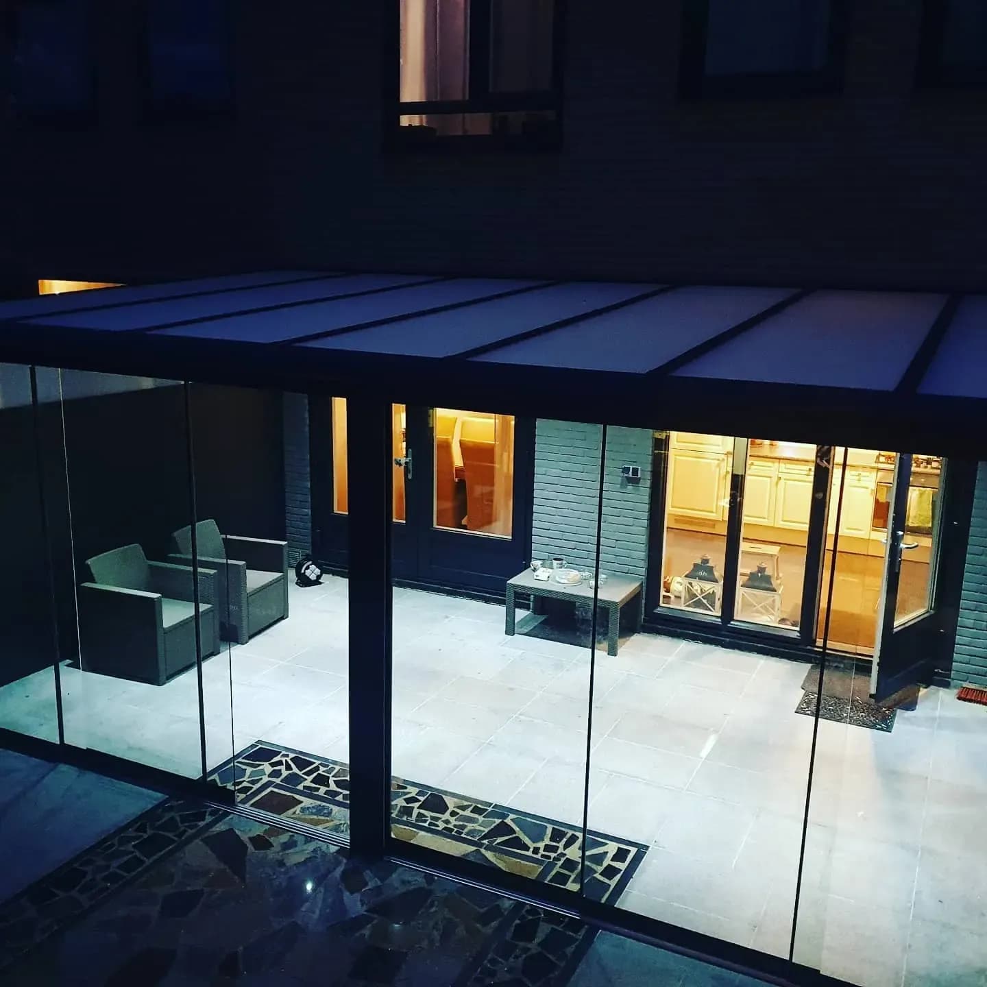 Glazen wanden van een veranda in de avond, met een zitgroep erachter en de verlichte woning op de achtergrond