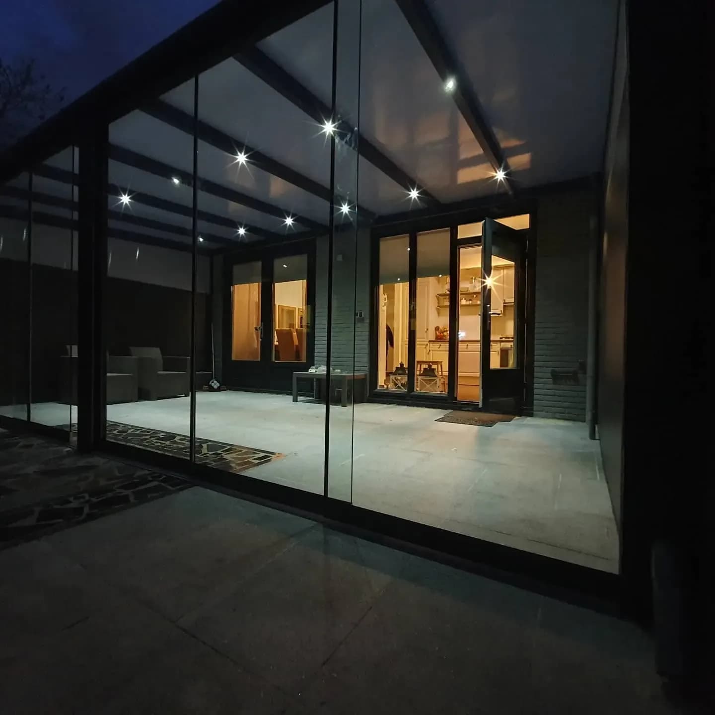 Overkapping in de avond met inbouwspots in het dak, glazen wanden en een verlicht huis erachter