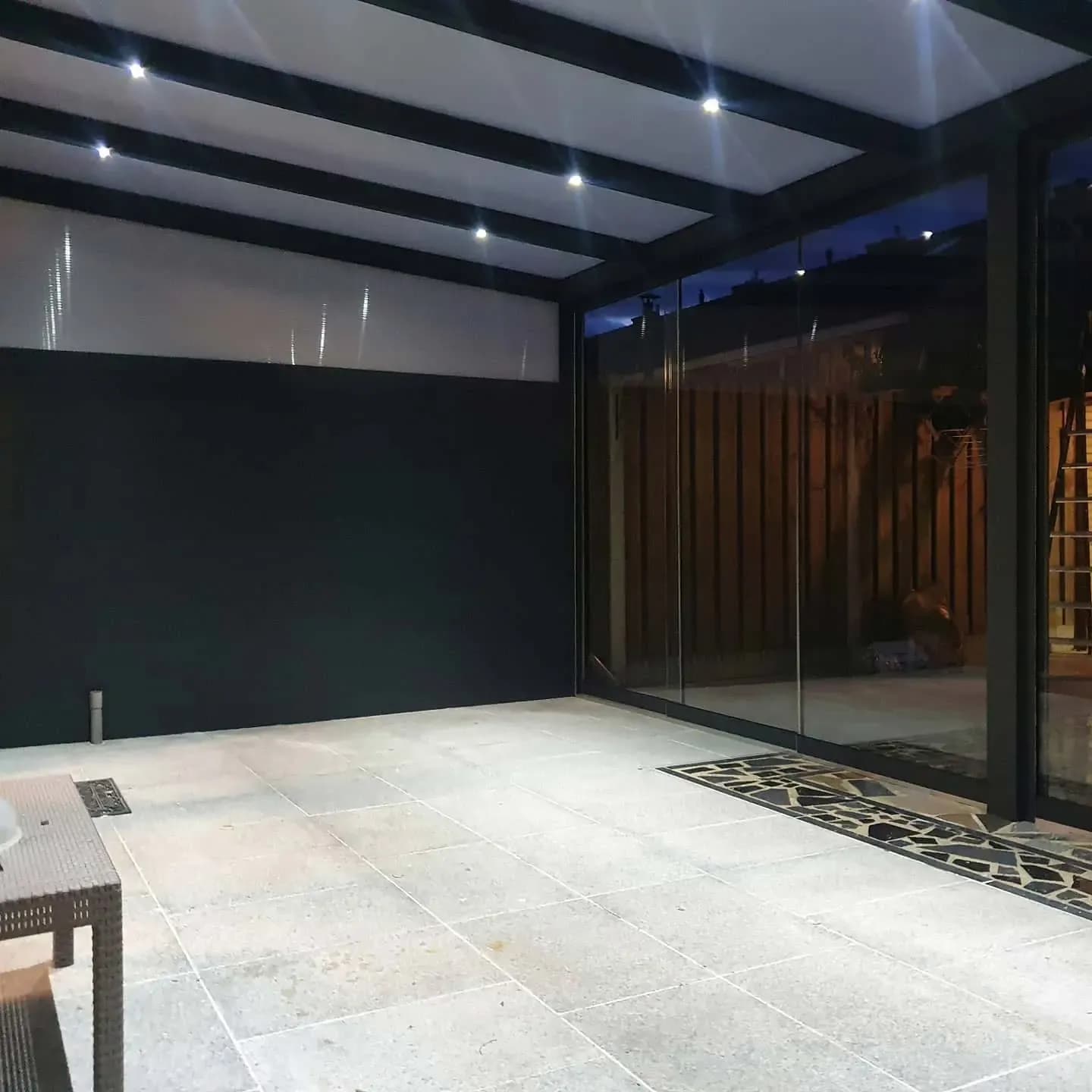 Overkapping in de avond met spots in het dak, een dichte wand en glas