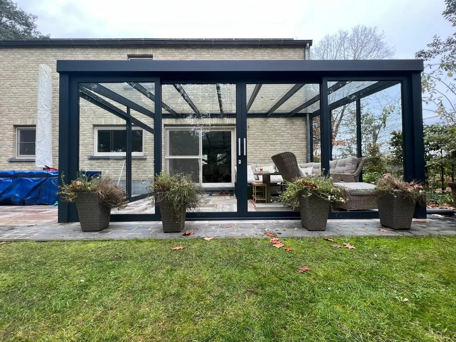 Veranda met glazen wanden en een lichtdoorlatend dak tegen een bakstenen woning, met plantenbakken op het terras