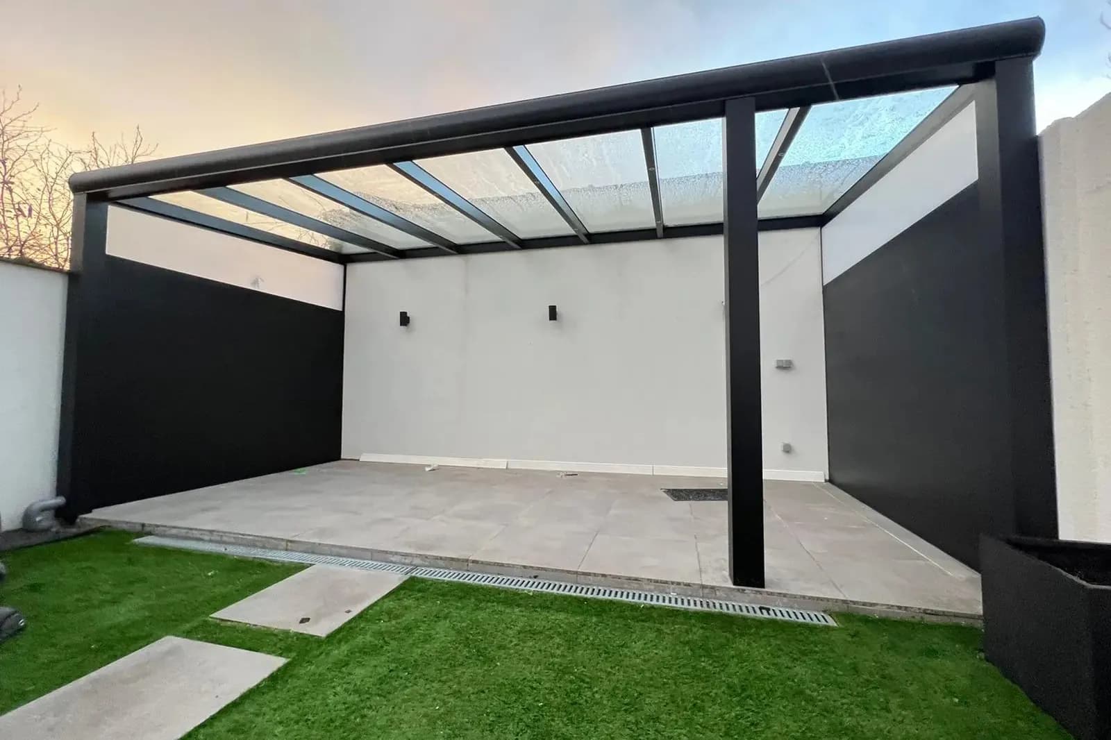 Veranda met een lichtdoorlatend dak, een witte achterwand en antracietkleurige zijwanden op een gazon, in avondlicht
