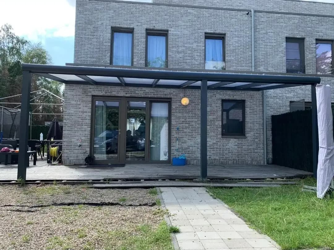 Brede veranda met een lichtdoorlatend dak tegen een grijs bakstenen huis, met een tuinpad ervoor