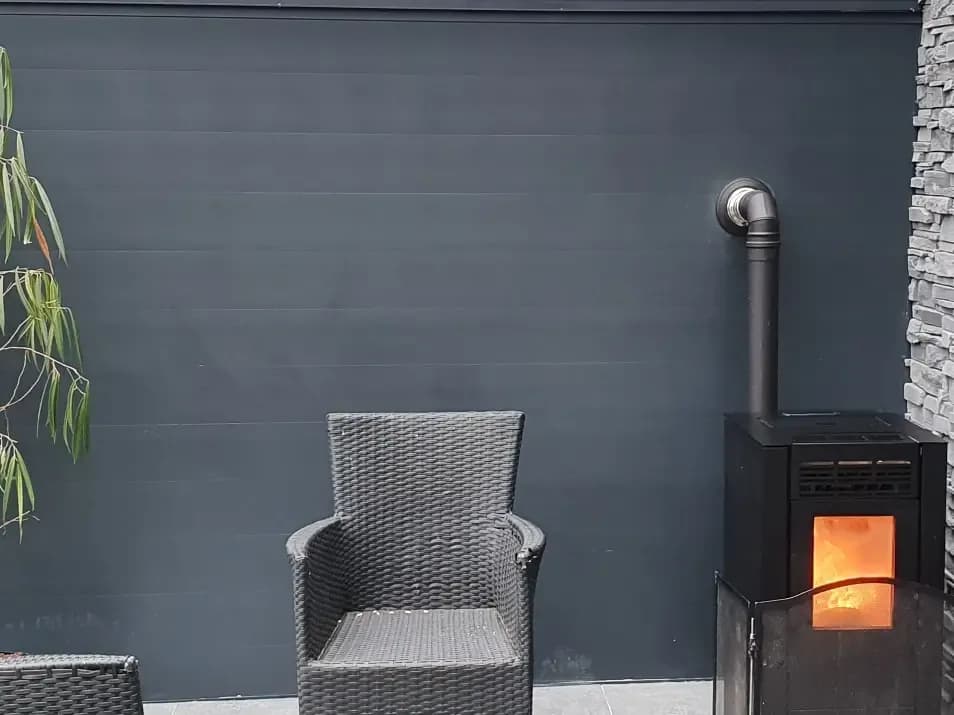 Antracietkleurige wand met een brandende kachel en een gevlochten tuinstoel ervoor